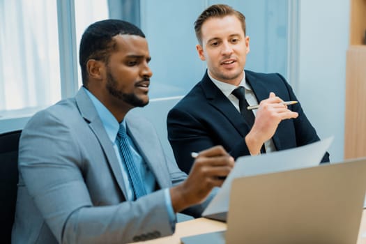 African male leader explain strategy while show solution to professional project manager by pointing at document, using laptop. Skilled business team brainstorming idea. Business meeting. Ornamented.