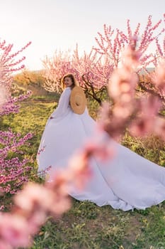 Woman blooming peach orchard. Against the backdrop of a picturesque peach orchard, a woman in a long white dress and hat enjoys a peaceful walk in the park, surrounded by the beauty of nature