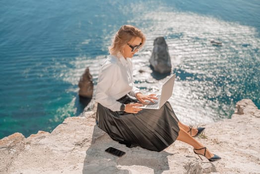 Business woman on nature in white shirt and black skirt. She works with an iPad in the open air with a beautiful view of the sea. The concept of remote work