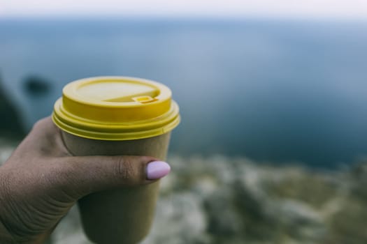 Hand holding Yellow cup with lid, coffee against a backdrop of a blue sky and sea. Illustrating cup and beverage.