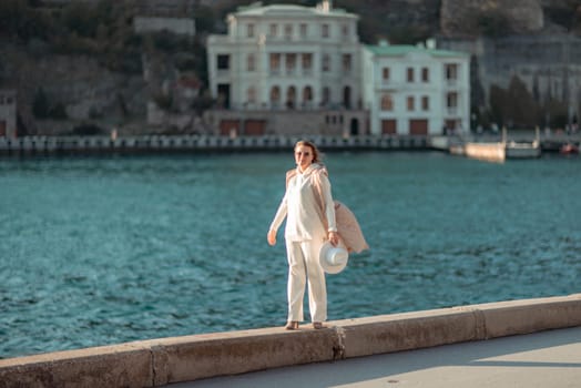 Happy blonde woman in a white suit and hat posing at the camera against the backdrop of the sea.