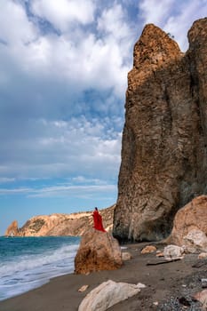 woman sea red dress. Woman with long hair on a sunny seashore in a red flowing dress, back view, silk fabric waving in the wind. Against the backdrop of the blue sky and mountains on the seashore