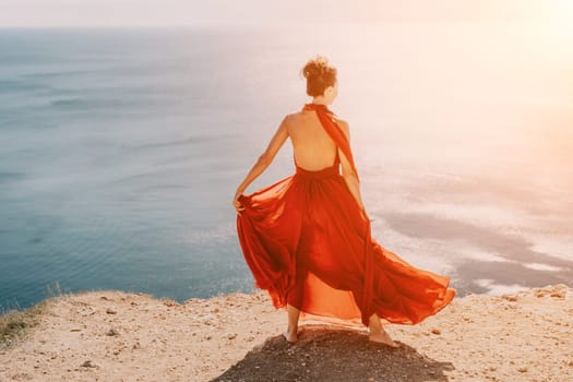 Side view a Young beautiful sensual woman in a red long dress posing on a rock high above the sea during sunrise. Girl on the nature on blue sky background. Fashion photo.