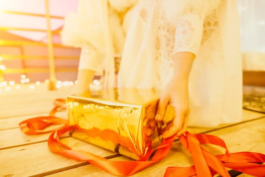 A woman in a white dress is holding a gold box with a red ribbon. She is wearing a crown on her head. The scene takes place in a room with a door and a window. The woman appears to be opening the gift box.