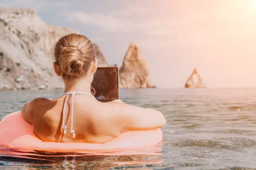 Woman freelancer works on laptop swimming in sea on pink inflatable ring. Pretty lady typing on computer while floating in the sea on inflatable donut at sunset. Freelance, remote work on vacation