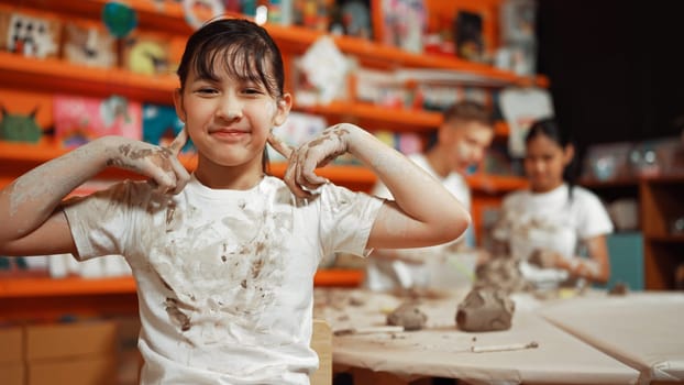 Asian girl pose at camera while diverse children modeling clay behind. Happy cute student wearing dirty shirt while looking at camera at workshop in art lesson. Blurring background. Edification.