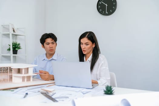 Professional male asian architect using ruler to measure house model length while young beautiful caucasian colleague using laptop to analyzed data on meeting table with house model. Immaculate.