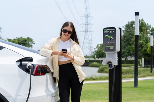 Young woman use smartphone to pay for electricity at public EV car charging station green city park. Modern environmental and sustainable urban lifestyle with EV vehicle. Expedient