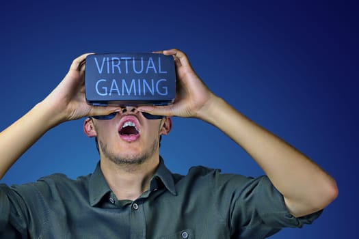 Young man in shirt enjoys and amazed by virtual reality glasses with virtual gaming inscription on them in front of blue background