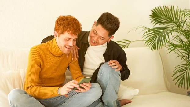 Gay couple smiling while using the mobile sitting on the sofa at home