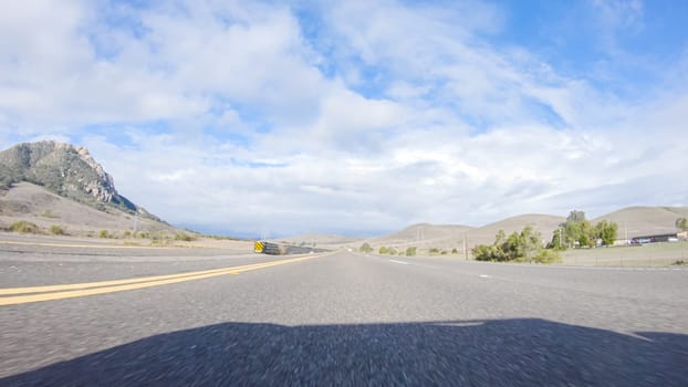 On a crisp winter day, a car cruises along the iconic Highway 101 near San Luis Obispo, California. The surrounding landscape is brownish and subdued, with rolling hills and patches of coastal vegetation flanking the winding road.