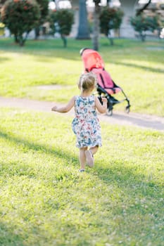 Little girl walks along a green lawn to a stroller standing on a path. Back view. High quality photo