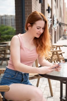 Young woman with braces on her teeth smiles while sitting in an outdoor cafe and uses a smartphone. Vertical photo.