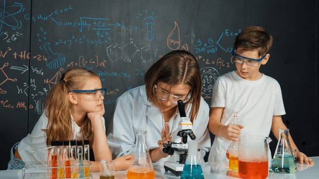 Smart teacher looking under microscope while teacher giving advice. Professional instructor wearing lab suit looking for diverse student at table with beaker filled with colored solution. Erudition.