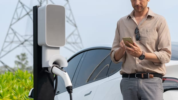 Man pay for electricity with smartphone while recharge EV car battery at charging station connected to power grid tower electrical as electrical industry for eco friendly car utilization.Expedient