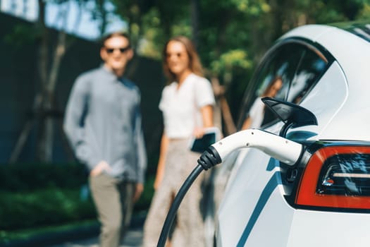 Young couple travel with EV electric car charging in green sustainable city outdoor garden in summer shows urban sustainability lifestyle by green clean rechargeable energy of electric vehicle innards