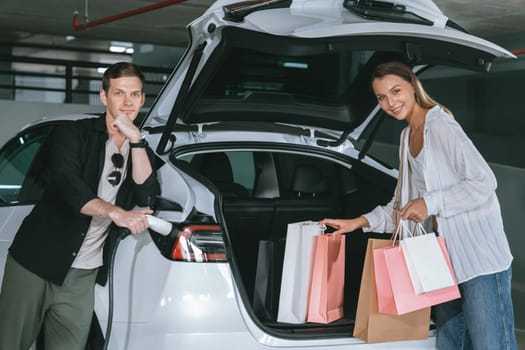 Young couple travel with EV electric car to shopping center parking lot charging in downtown city showing urban sustainability lifestyle by green clean rechargeable energy of electric vehicle innards