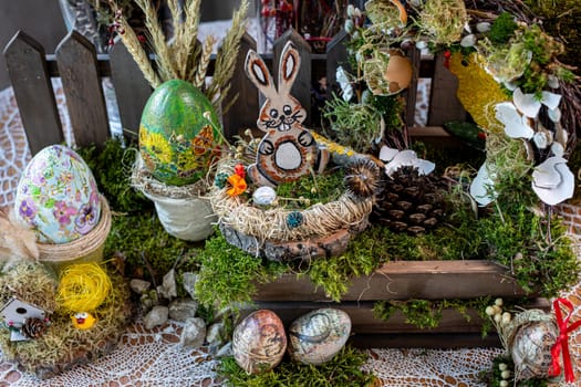 The scene captures a charming Easter composition viewed from above, featuring a wooden rabbit figurine nestled in a bird's nest atop a wooden crate. In the background, colorful Easter eggs displayed in pots and a wooden fence add to the rustic charm.