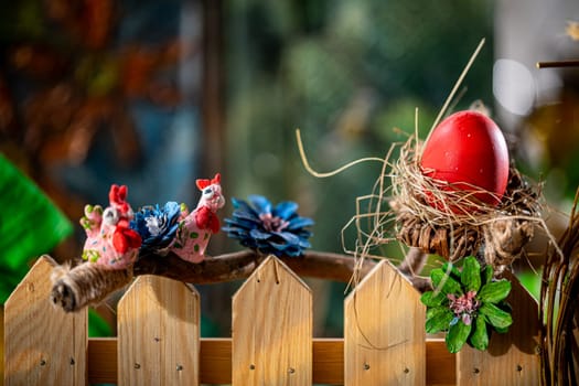 Amidst a wooden fence and arranged branches lies a nest intricately woven from dry straw, cradling a large egg painted in vibrant red.
