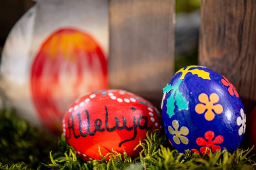 In the foreground of the image, two handcrafted Easter eggs steal the spotlight. One egg features a deep navy blue hue adorned with intricate floral patterns, while the other boasts a vibrant red color with the word ,Alleluja, delicately written upon it.
