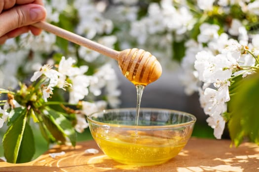 Fresh golden honey pours from a wooden dipper into a glass bowl, with blooming white flowers in the background.