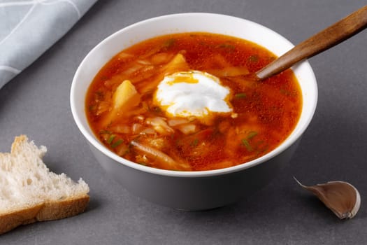 Close up borscht with sour cream in a bowl with bread on a gray table.