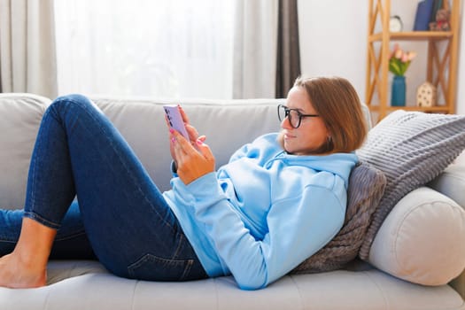 Smiling woman in glasses lounging comfortably and browsing on her phone at home