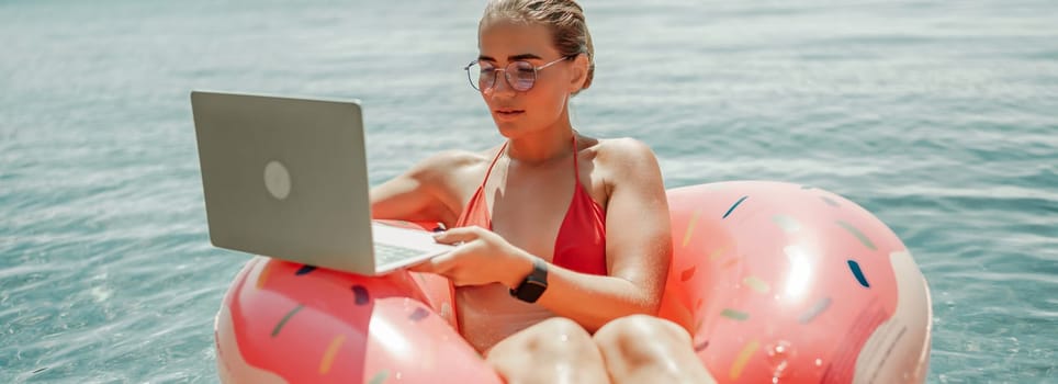 Woman laptop sea. Freelancer woman in sunglases floating on an inflatable big pink donut with a laptop in the sea. People summer vacation rest lifestyle concept