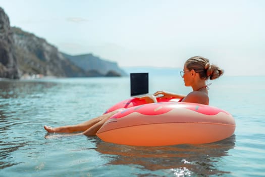 Woman laptop sea. Freelancer woman in sunglases floating on an inflatable big pink donut with a laptop in the sea. People summer vacation rest lifestyle concept