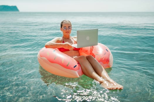 Woman laptop sea. Freelancer woman in sunglases floating on an inflatable big pink donut with a laptop in the sea. People summer vacation rest lifestyle concept