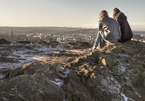 Edinburgh, Scotland - Jan 17, 2024 - Two young women sitting relaxing on top of rock over the mountain and enjoying looking at scenic view of cityscape. Two female Sitting down enjoying the amazing view from the mountain top of Salisbury Crags, Destinations in Europe, Space for text, Selective focus.