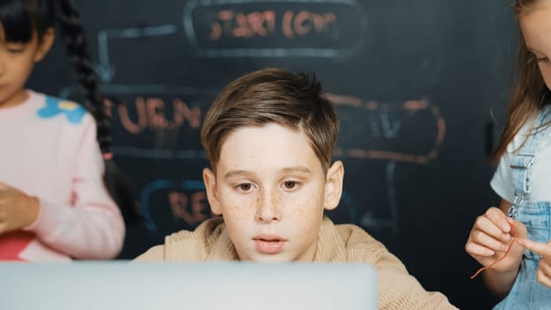 Closeup of boy using laptop programing engineering code and writing program while group of diverse kid holding controller in STEM technology classroom at blackboard written with prompt. Erudition.