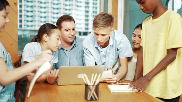 Caucasian teacher giving advise to young student while boy lecture comment. Professional instructor write comment surrounded by multicultural children at table with laptop and paper. Edification.