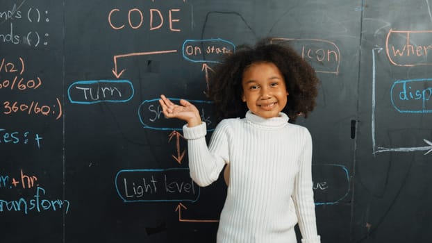 Happy african girl writing blackboard and turnaround to waving at camera while standing at board with engineering prompt or coding, programing system written in STEM technology classroom. Erudition.