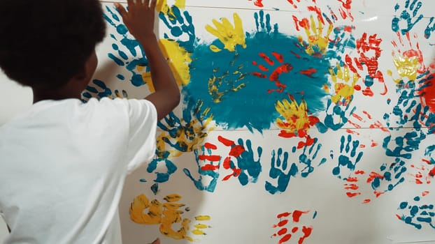 Back view of happy african boy painted the stained wall with colorful hand while wearing casual white shirt in art lesson.Smart student use hand print to make creative artwork. Education. Edification.