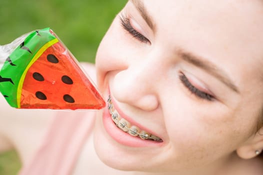 Beautiful young woman with braces on her teeth eats a watermelon-shaped lollipop outdoors