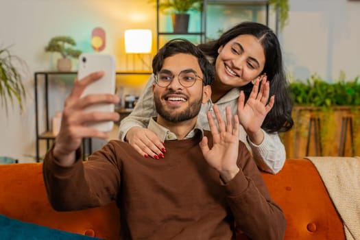 Diverse happy Hispanic couple waving hello while talking on smartphone video call on sofa in room at home. Cheerful girlfriend and boyfriend in casual clothes recording vlog on cellphone in apartment.