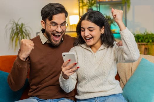 Happy Arabian couple celebrating success win reading great news on smartphone while sitting on sofa in living room at home. Excited smiling Indian family using cellphone together clenching fists.