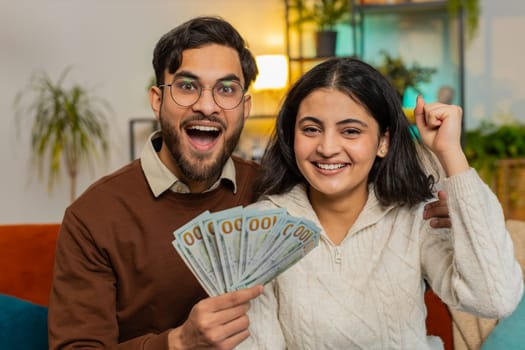 Happy young Indian couple holding showing money cash banknote together looking at camera sits on home sofa in room. Successful rich Hispanic family clenching fists celebrate lottery jackpot game win