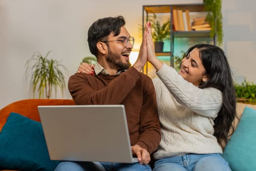 Cheerful young diverse couple in casual clothes using laptop and surprised with email winning lottery on sofa in living room at home. Happy Indian family giving high-five clenching fists in apartment.