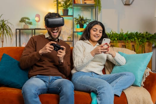 Happy young man wearing VR goggles playing videogame with joystick sitting beside Indian girlfriend using smartphone sitting on sofa in living room at home. Happy family spending weekend in apartment.
