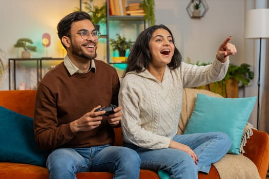 Cheerful young Indian couple using joystick controller playing video game fun enjoying sitting on sofa in living room. Diverse family enjoying success winning online game during weekend in house