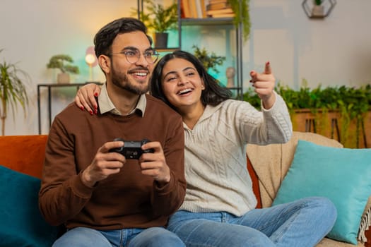 Cheerful young Hispanic couple using joystick controller playing video game fun enjoying sitting on sofa in living room. Diverse family enjoying success winning online game during weekend in house