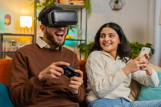 Happy exited man wearing VR goggles playing videogame with joystick sitting beside Indian girlfriend using smartphone sitting on sofa in living room at home. Happy family spending weekend in apartment