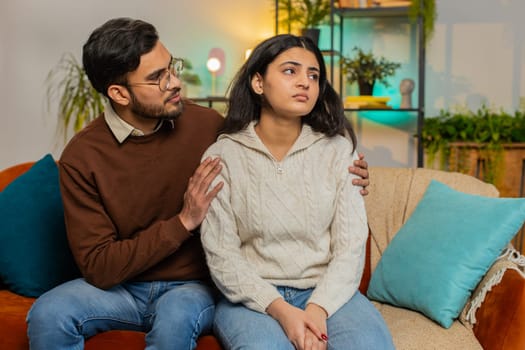 Compassion. Caring young man comforting upset Indian girlfriend sitting on sofa in room at home. Husband and wife family marriage couple together on couch in apartment. Man supporting depressed woman.