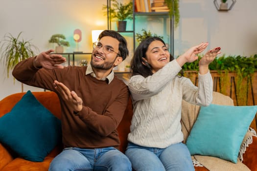 Excited diverse rich couple wasting or throwing money hand gesture sitting on sofa in living room at home. Successful Indian family giving tips earnings, big profit, win lottery, celebrate concepts.