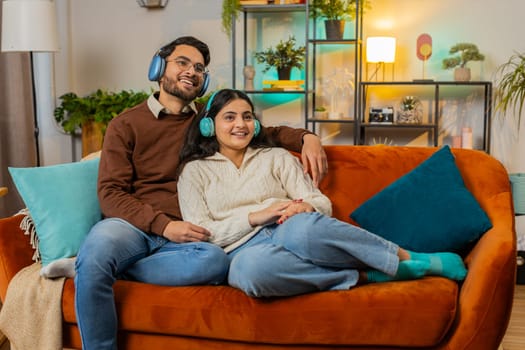 Cheerful happy young diverse couple in wireless headphones dancing on couch at home listening favorite energetic disco rock n roll music. Smiling relaxed family enjoying weekend together in apartment.