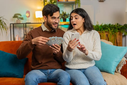 Happy Indian couple holding money cash and smartphone planning discussing investment together sitting on sofa in living room at home. Successful exited amazed diverse family on couch in apartment.
