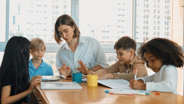 Professional caucasian teacher telling story to diverse student while sitting at table with storybook and colored book. Smart learner listening story while colored picture from instructor. Erudition.