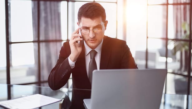 Portrait of a young serious businessman in a casual office calling on the phone while working with a laptop